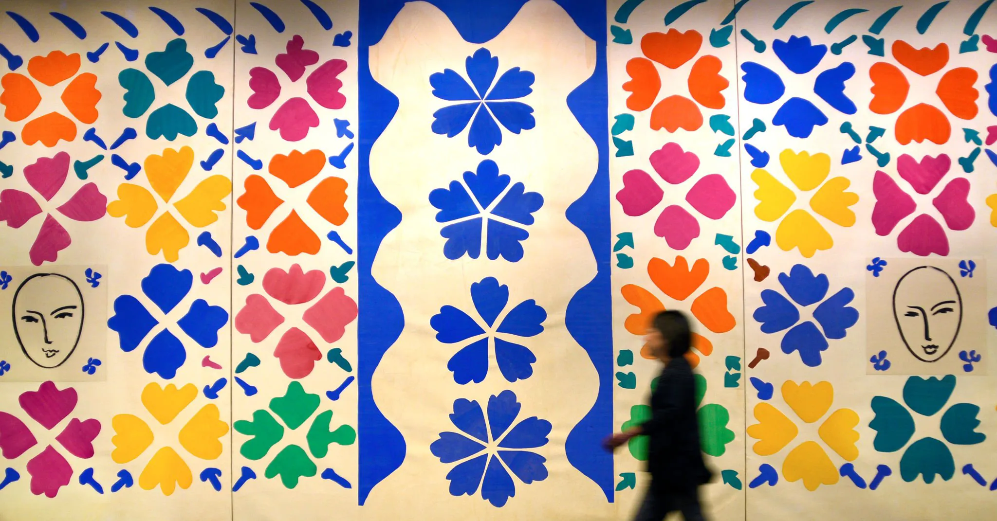 A museum wall hung with cut-out floral panels in blue, orange, pink and yellow, a visitor walking past.